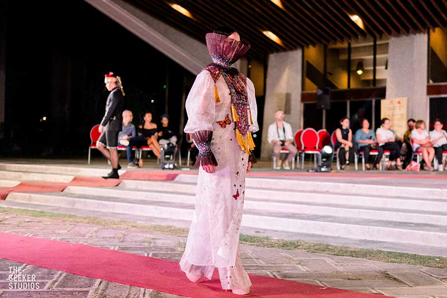 Model in a butterfly-embellished white gown with intricate layered detailing and bold accessories