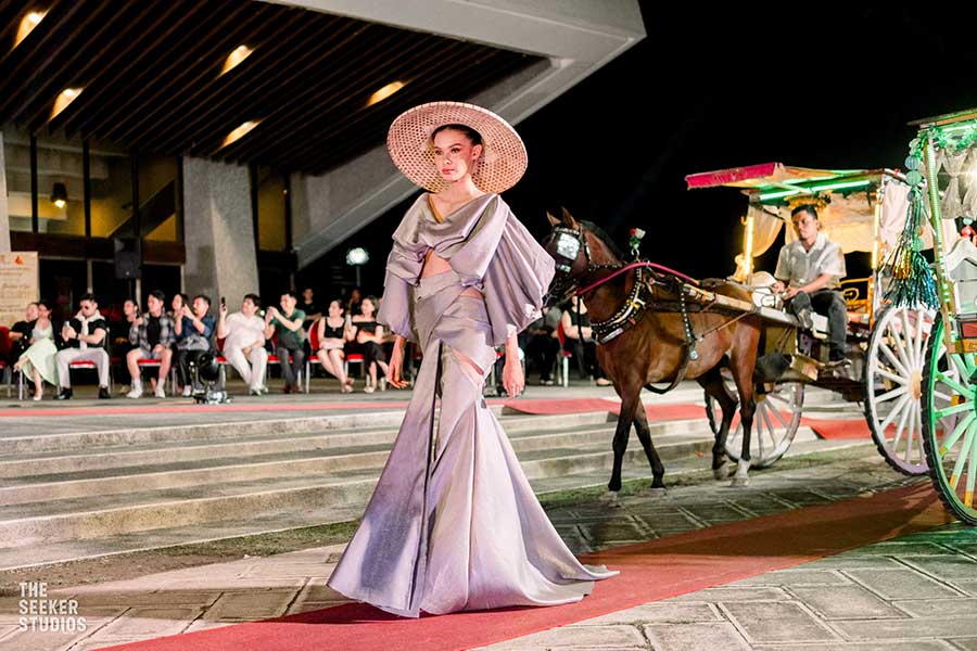 model in a lavender gown and straw hat walking near horse-drawn carriage