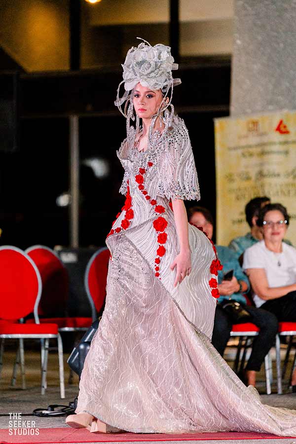 Model in a silver gown with intricate red rose embellishments and a dramatic sculpted headpiece