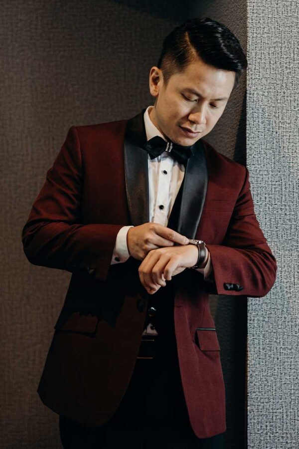 Man in a sophisticated burgundy suit with black satin lapels, adjusting his watch