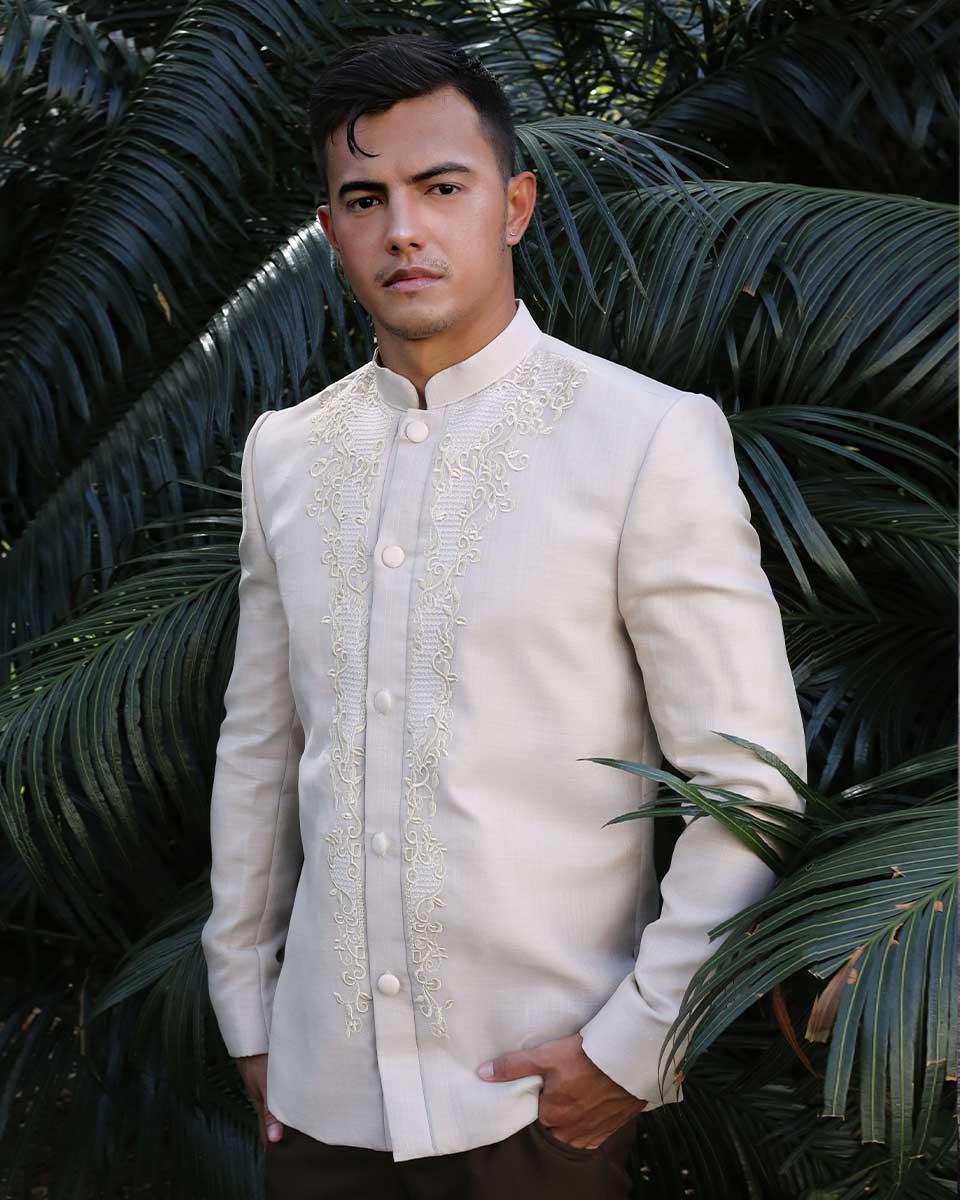 A man wearing a sophisticated Coat Barong with intricate embroidery, standing against lush green palm leaves in an elegant outdoor setting