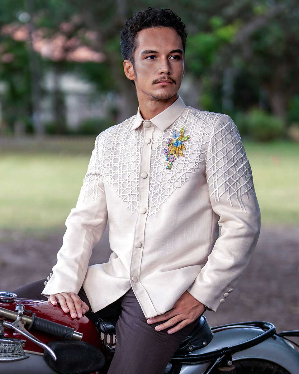 A man wearing a stylish Modern Barong with geometric embroidery and a brooch, sitting on a vintage motorcycle in an outdoor setting