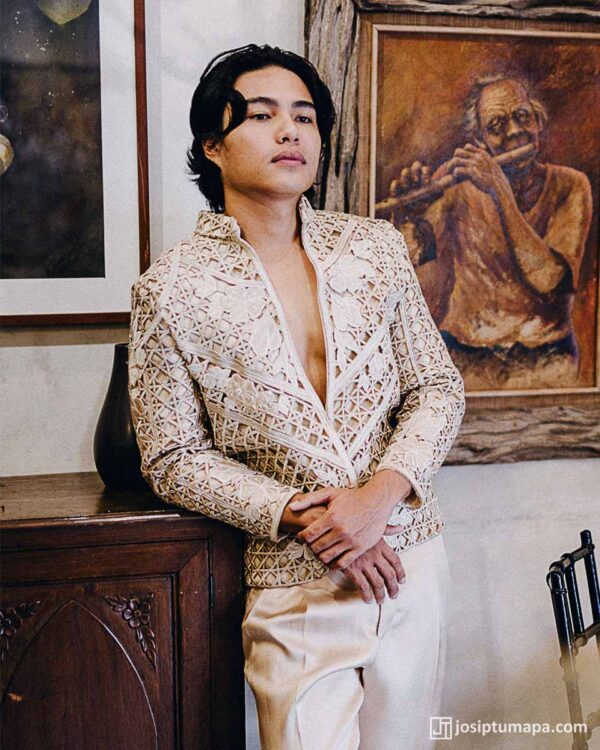Man in avant-garde ivory Barong Tagalog with lattice embroidery, leaning on a wooden table, rustic painting in the background