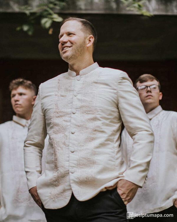 Groom in cream Barong Tagalog at outdoor wedding with groomsmen, stone wall, and greenery