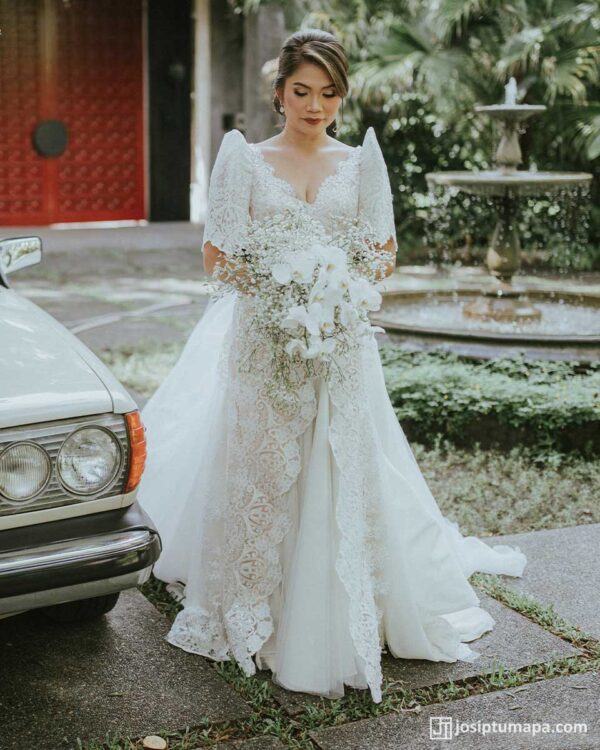 Filipiniana wedding gown with lace and butterfly sleeves, paired with a white orchid bouquet near a vintage car and fountain