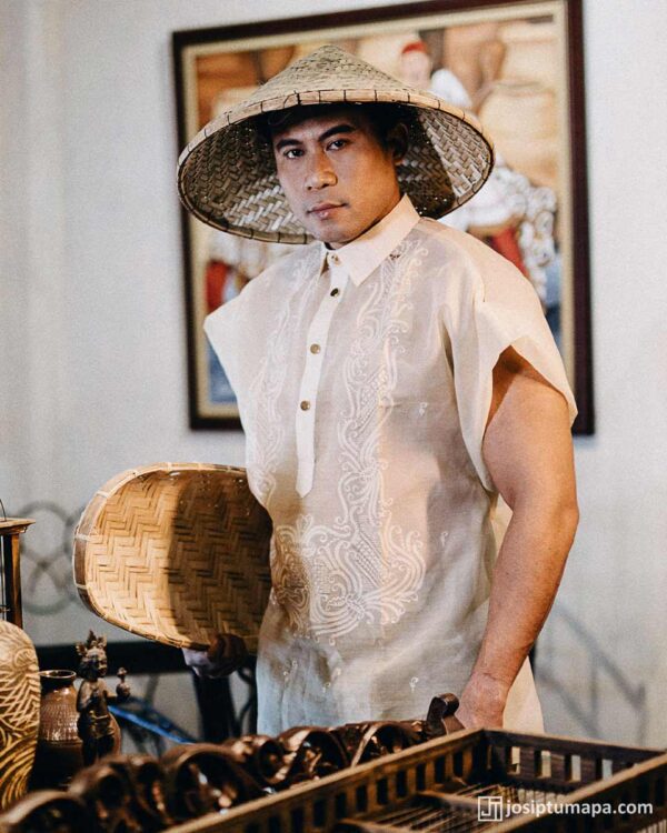 Man in cream short-sleeve Barong Tagalog with white embroidery, conical hat, and woven basket, cultural painting behind