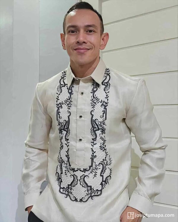 Man in cream Piña Jusi Barong Tagalog with black embroidery, against a slatted wall