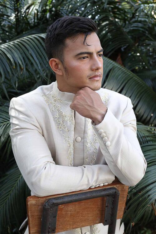 Man in cream Barong Tagalog coat with embroidery, leaning on a chair with palm leaves background