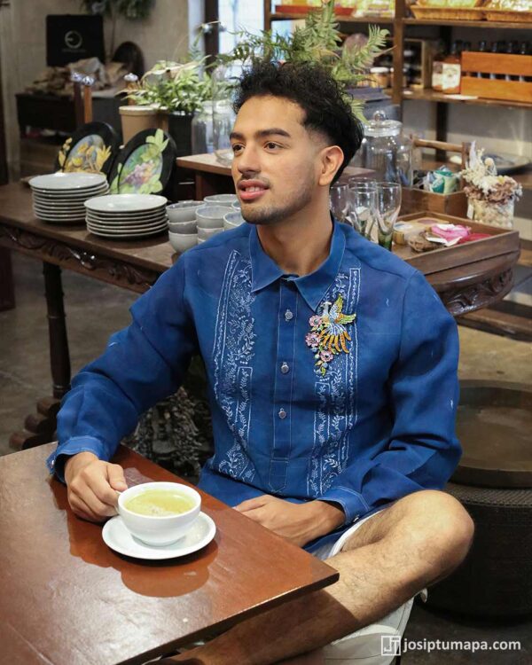 Man wearing deep-blue cocoon silk Barong Tagalog with floral brooch accent, seated in a café setting