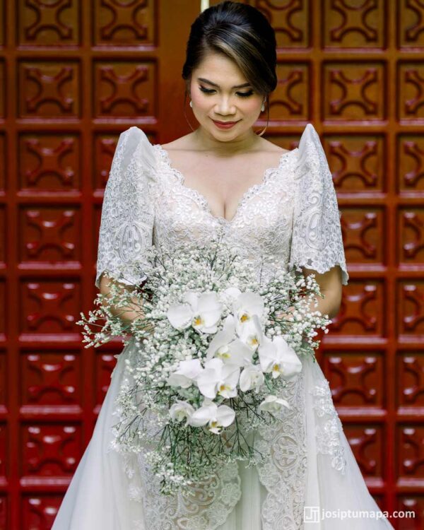 Bride in handcrafted Filipiniana wedding gown with lace embroidery and butterfly sleeves holding white orchid bouquet