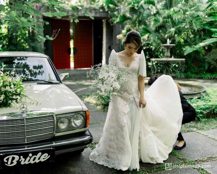 Bride in Filipiniana lace wedding gown walking in garden with vintage car and white orchid bouquet
