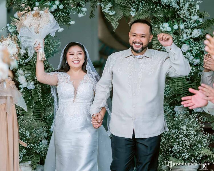 Filipino wedding couple celebrating under floral arch — groom in bespoke Piña Cocoon Silk Coat Barong, bride in handcrafted lace gown