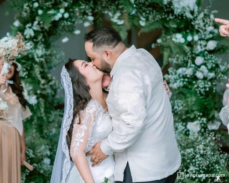 Groom wearing custom Piña Cocoon Silk Barong kisses bride during Filipino wedding ceremony under floral arch