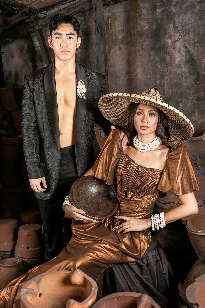 Man in a dark tailored jacket and woman in a bronze terno-inspired gown with a wide hat and layered necklace