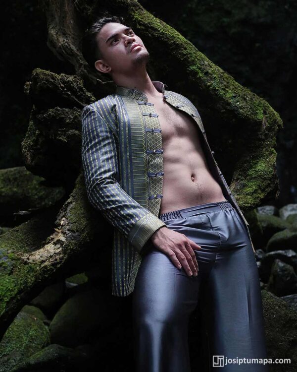 Model leaning on moss-covered roots, patterned jacket and silver trousers in dark forest