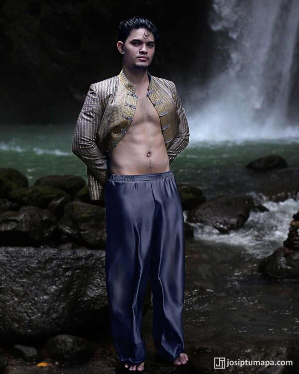 Model barefoot on rocks before waterfall, open patterned jacket and grey trousers