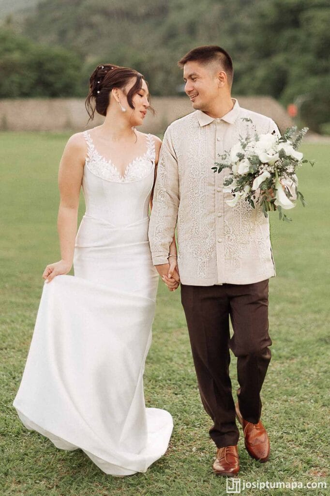 Groom in an embroidered wedding Barong Tagalog walking with a bride in a white gown during an outdoor ceremony
