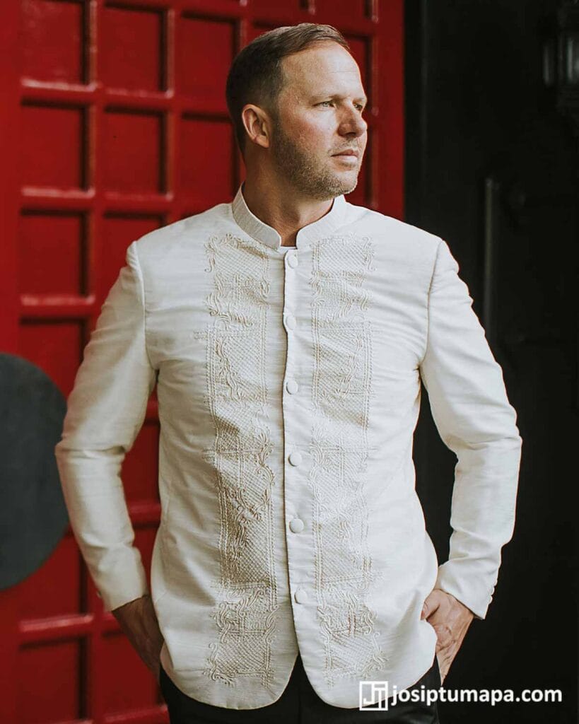 Man wearing a white coat-style Barong Tagalog with tonal embroidery against a red architectural background